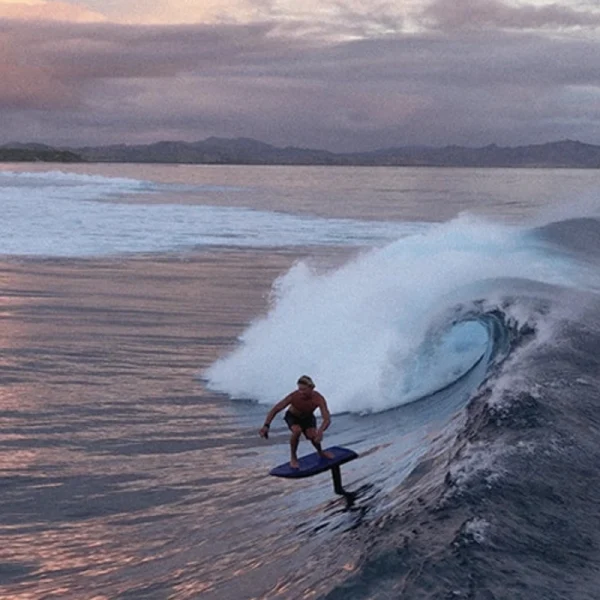 Fliteboard Ultra L3 - Lifestyle Image 6 A surfer riding a Fliteboard Ultra L3 efoil on a breaking ocean wave at sunset.